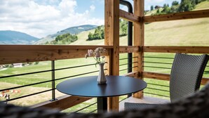 A wooden balcony with a round table and a wicker chair, overlooking a green landscape with hills and a clear sky. - Mareo Dolomites Hotel (Marebbe)