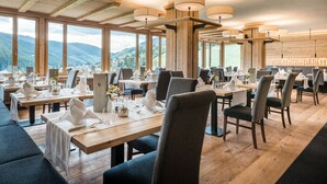 A restaurant with wooden tables and chairs, large windows offering mountain views, and hanging pendant lights. - Mareo Dolomites Hotel (Marebbe)