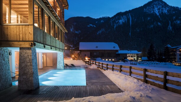 A wooden building with a stone foundation, a wooden deck, and a snow-covered landscape with mountains in the background. - Mareo Dolomites Hotel (Marebbe)