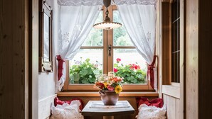A wooden interior with a window, lace curtains, a table with a vase of flowers, and two upholstered chairs. - Mareo Dolomites Hotel (Marebbe)