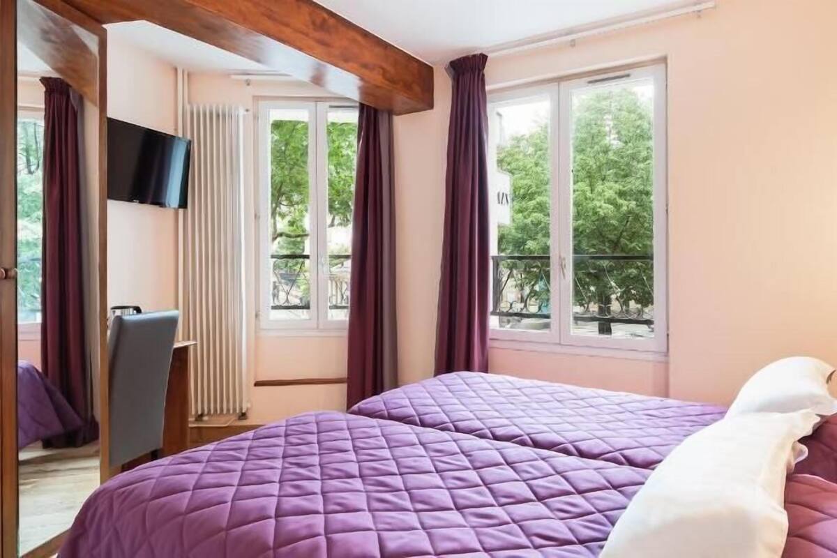 Et hotelværelse med to senge, et fjernsyn og store vinduer med gardiner.