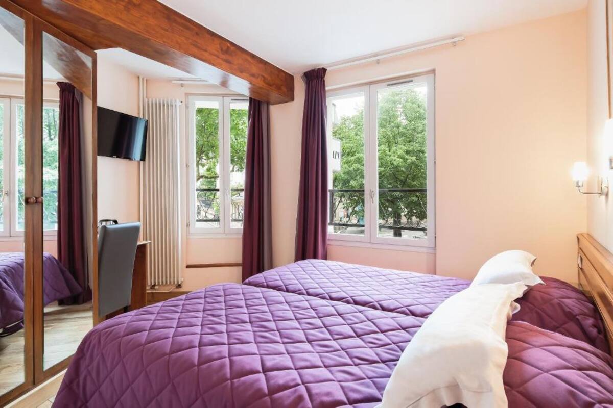 Et hotelværelse med to senge, et fjernsyn og en balkon med udsigt til træer.