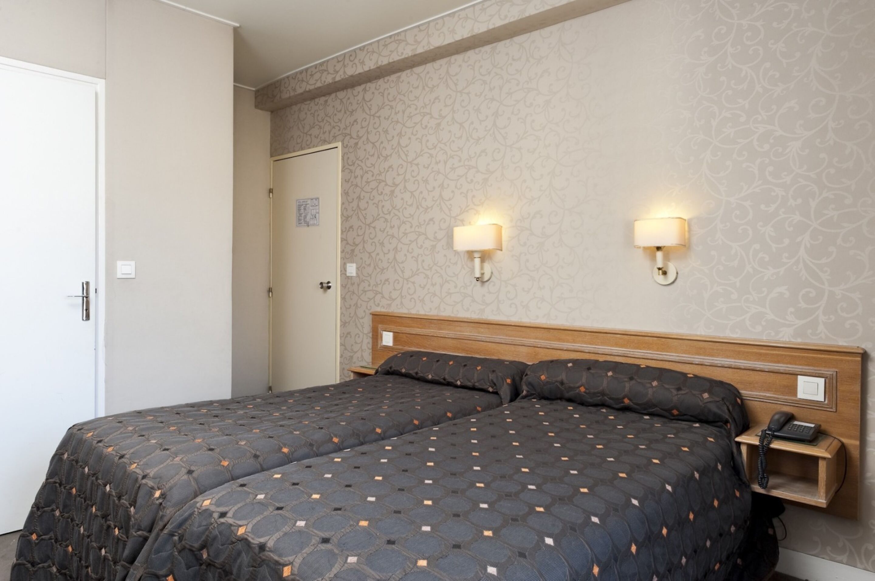 A hotel room with a double bed, two bedside tables with lamps, a wooden headboard, and a patterned wallpaper.