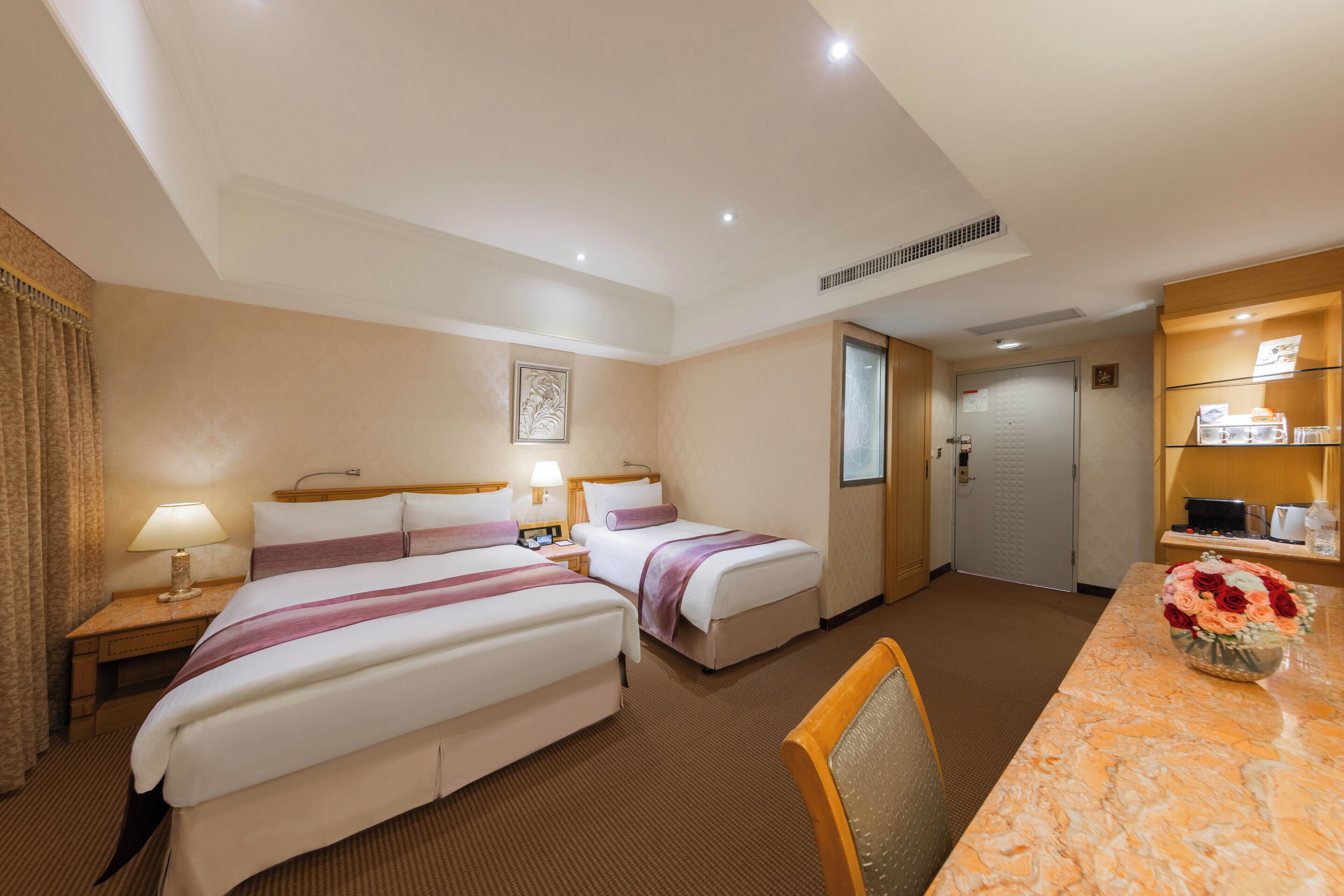 A hotel room with two beds, a desk, a chair, a bathroom, and a reception area.