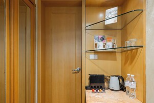 A hotel room with a wooden door, a glass shelf with mugs and a kettle, and a coffee maker on the counter.