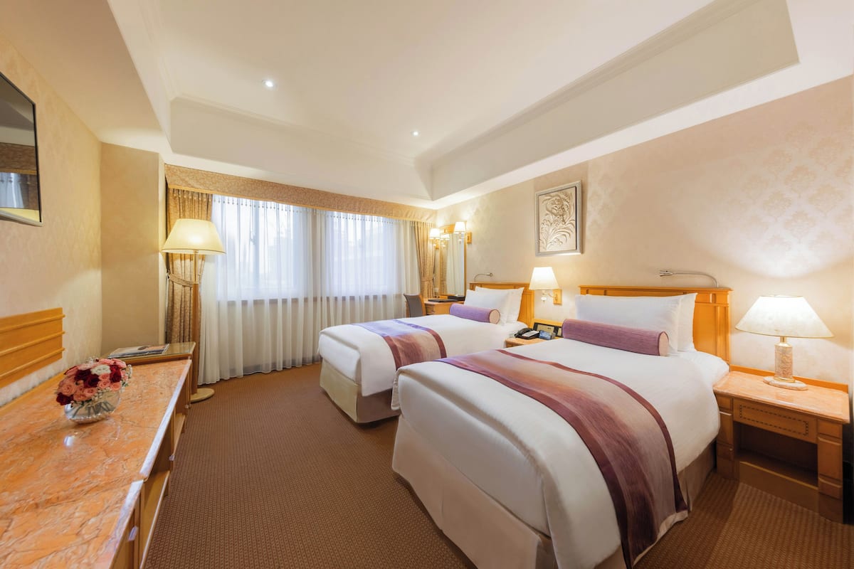 A hotel room with two beds, a desk with a vase of flowers, and a large window with curtains.