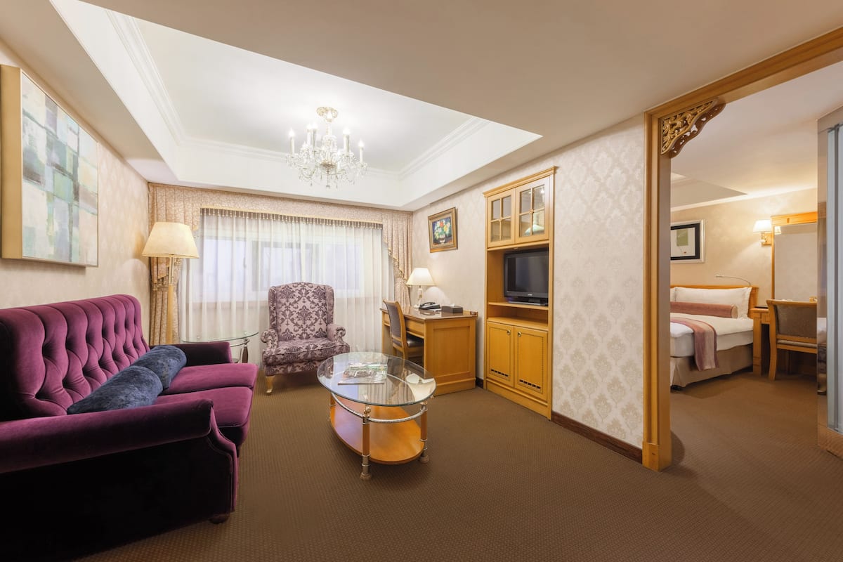 A hotel room with a plush purple sofa, a glass coffee table, a wooden cabinet with a television, and a large mirror.