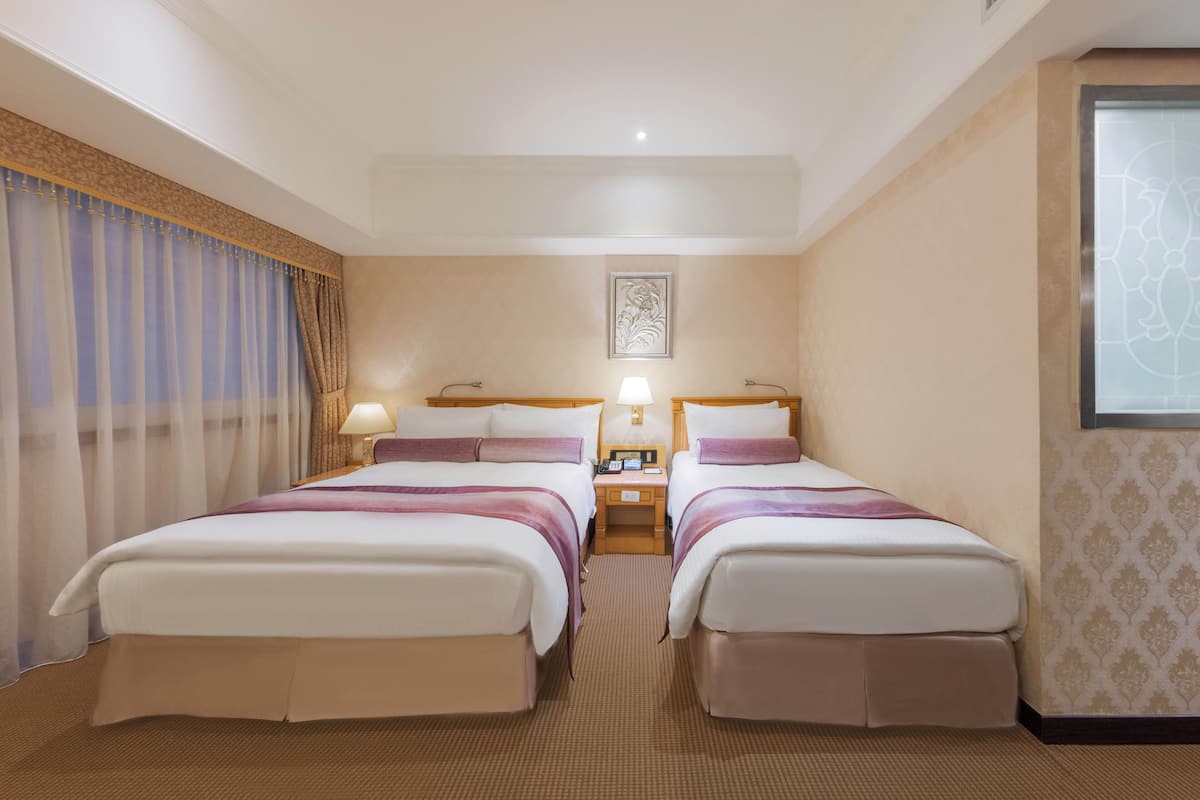 A hotel room with two beds, a nightstand with lamps, a framed artwork on the wall, and a window with a patterned curtain.