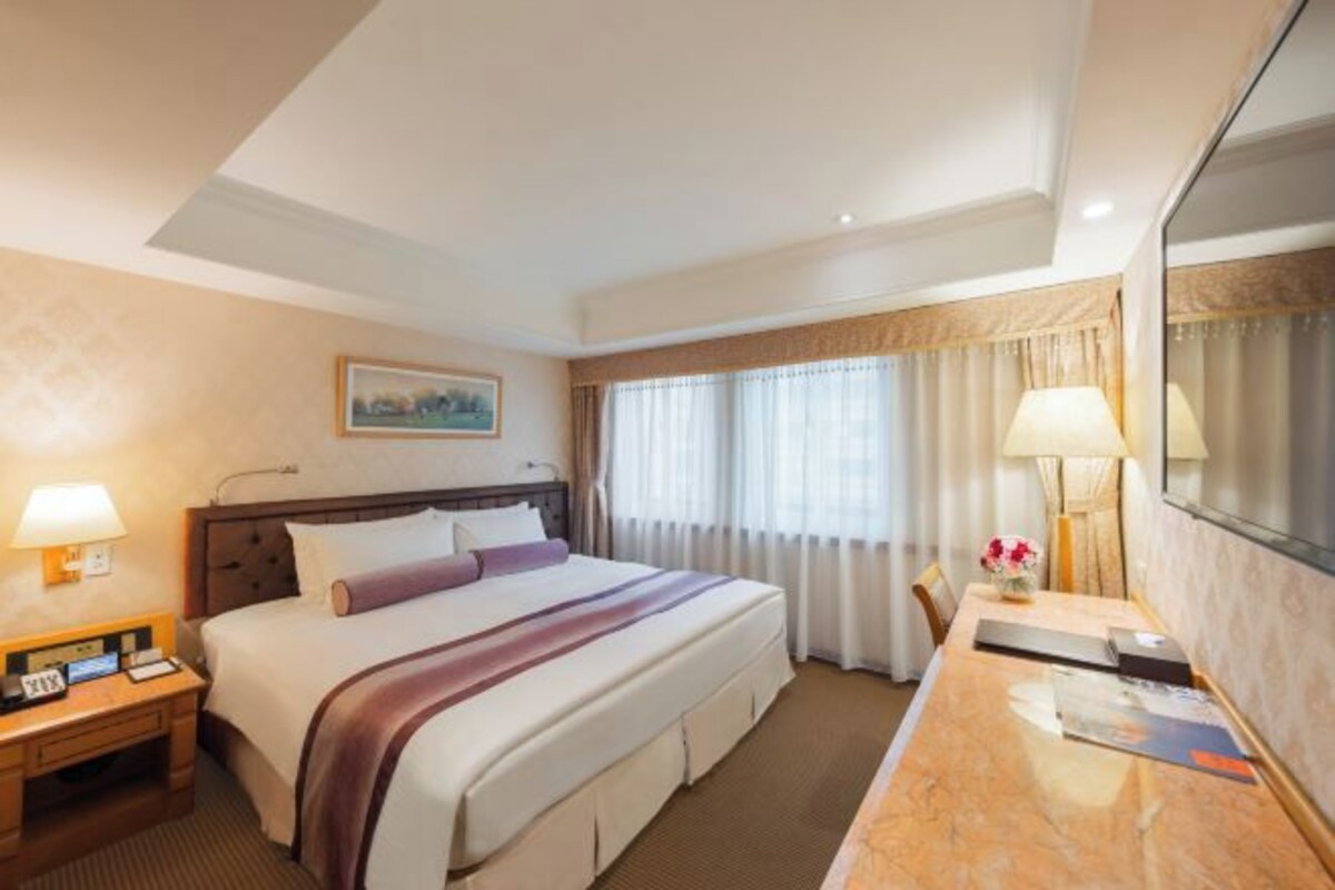 A hotel room with a large bed, a desk with a lamp, a chair, and a window with curtains.