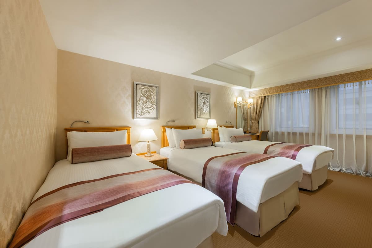 A hotel room with three beds, each with a headboard, bedside tables, lamps, and a large window with curtains.