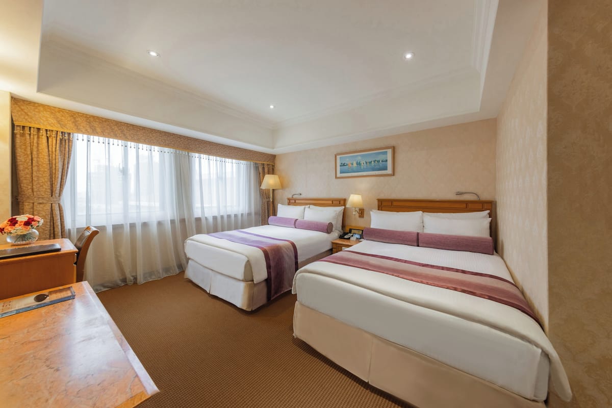 A hotel room with two beds, a desk, and a chair. There is a painting on the wall and a window with curtains.