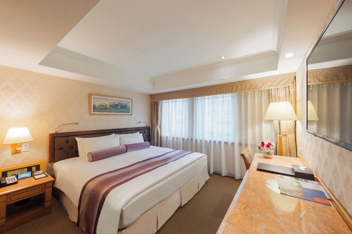 A hotel room with a large bed, a desk with a lamp, a mirror, and a window with curtains.