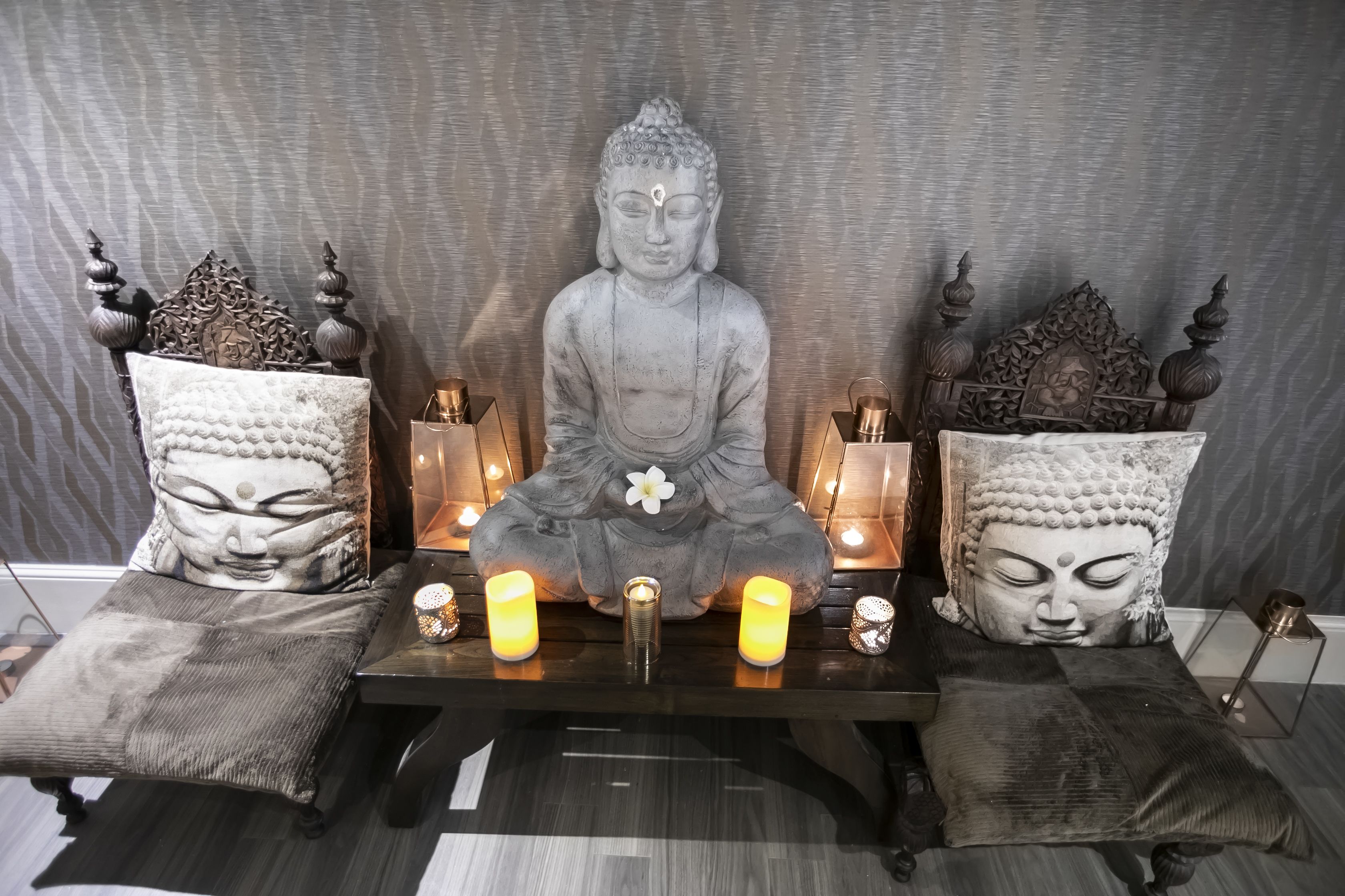 Eine Statue einer sitzenden Figur mit einer Blume auf der Brust, begleitet von zwei gleich designten Kissen und drei brennenden Kerzen auf einem Tisch davor.