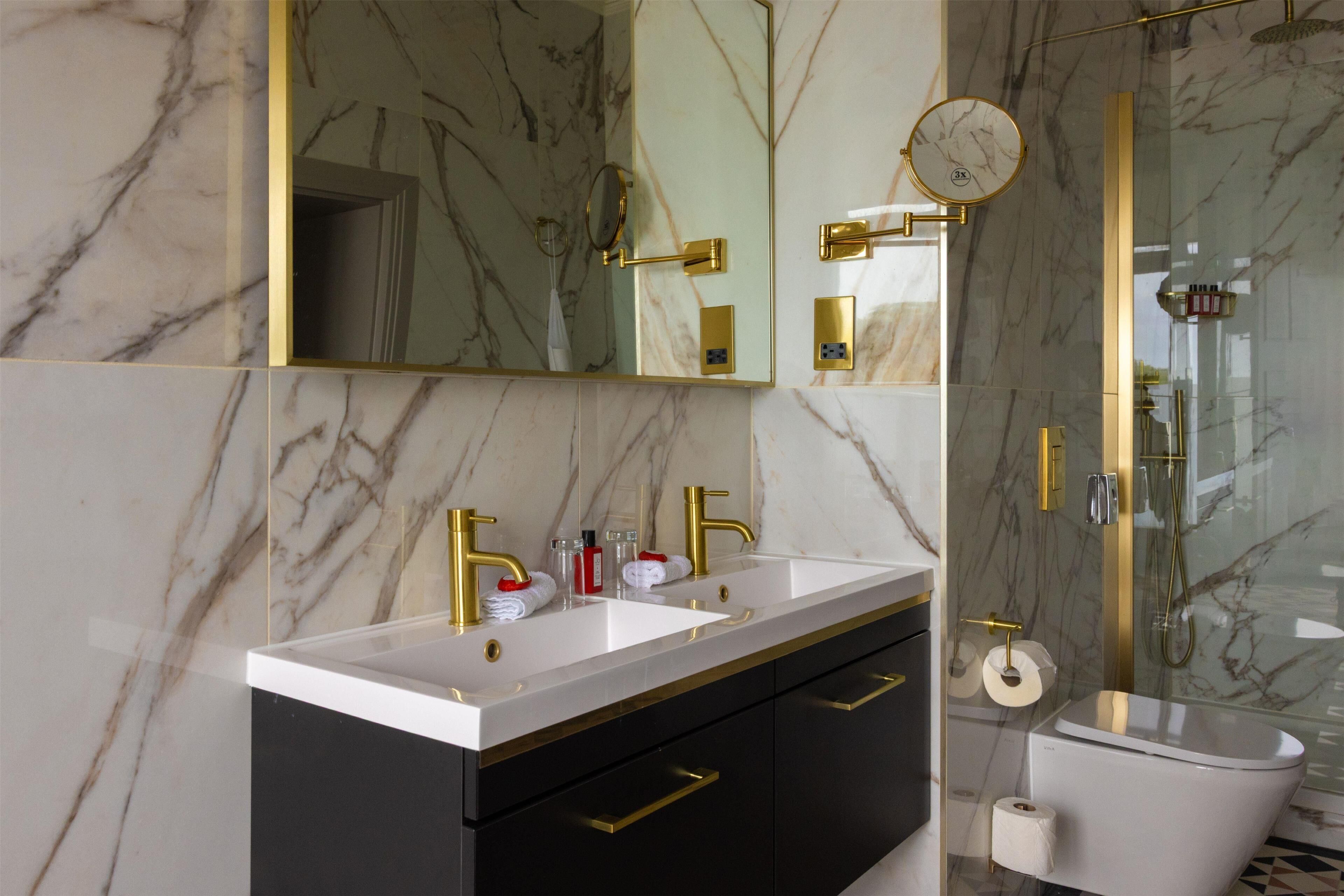 Une salle de bain aux murs de marbre, dotée d’un double lavabo avec des robinets dorés, de toilettes et d’une douche séparée par des parois en verre.
