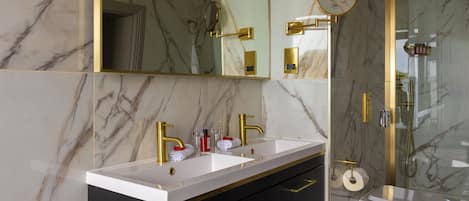 Une salle de bain aux murs de marbre, dotée d’un double lavabo avec des robinets dorés, de toilettes et d’une douche séparée par des parois en verre.