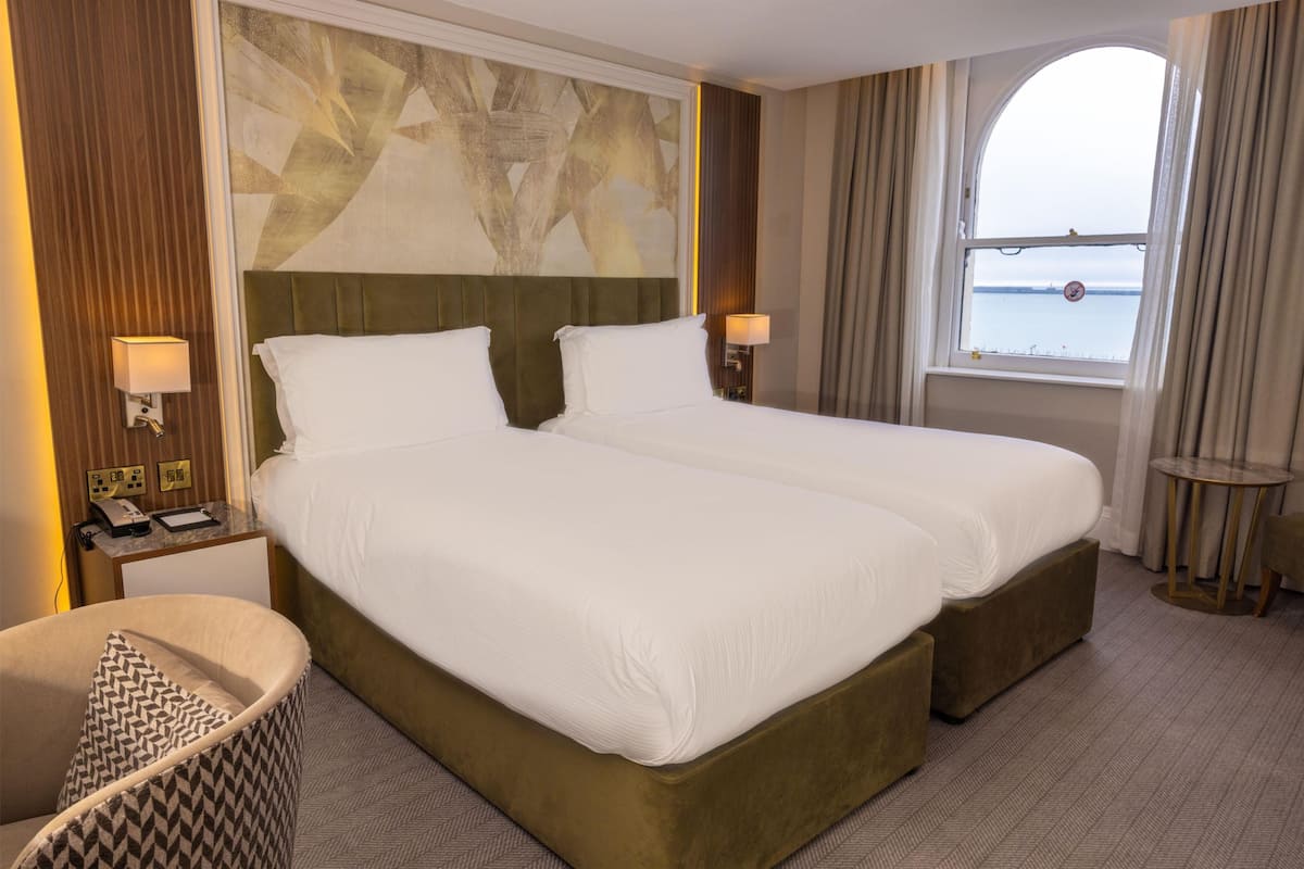 A hotel room with two beds, a view of the sea, a patterned chair, and a small table with a lamp.