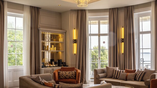 Een ruime slaapkamer met een groot bed, een sofa en een salontafel. Er hangt een kroonluchter aan het plafond en er is uitzicht op het groen door grote ramen.