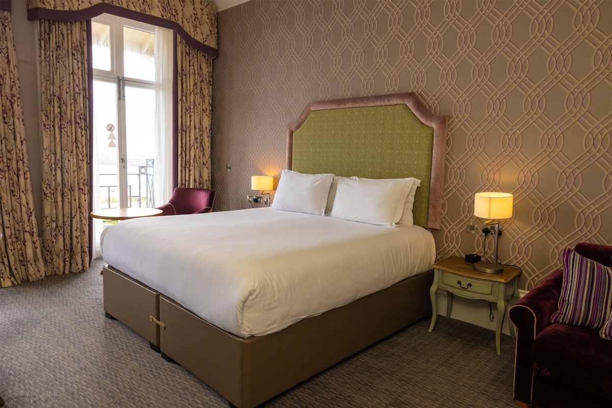 A hotel room with a large bed, two bedside tables with lamps, a chair, and a patterned wall.