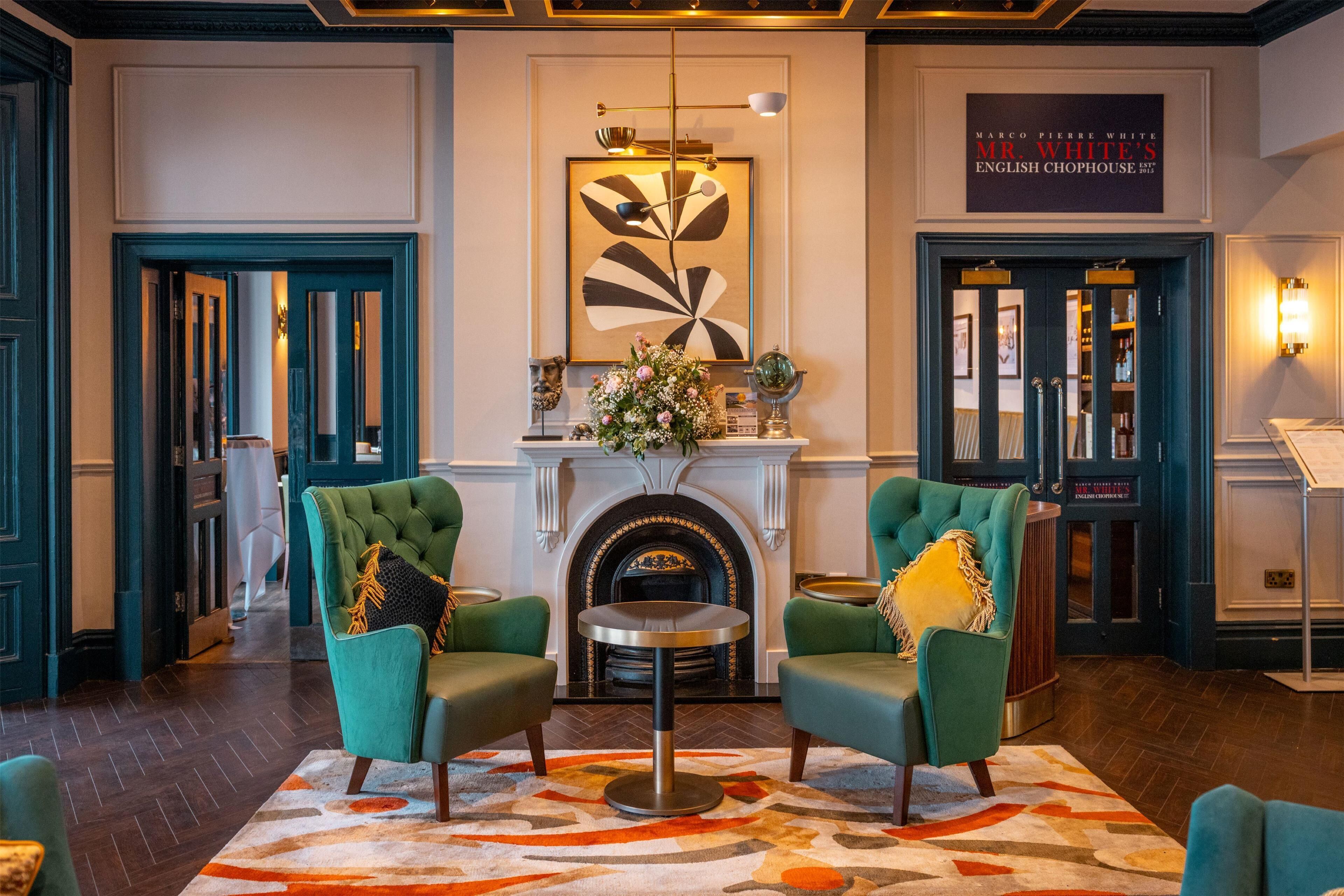 A lobby with two green armchairs, a round table, a fireplace, and a framed artwork above.