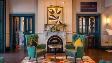 A lobby with two green armchairs, a round table, a fireplace, and a framed artwork above.
