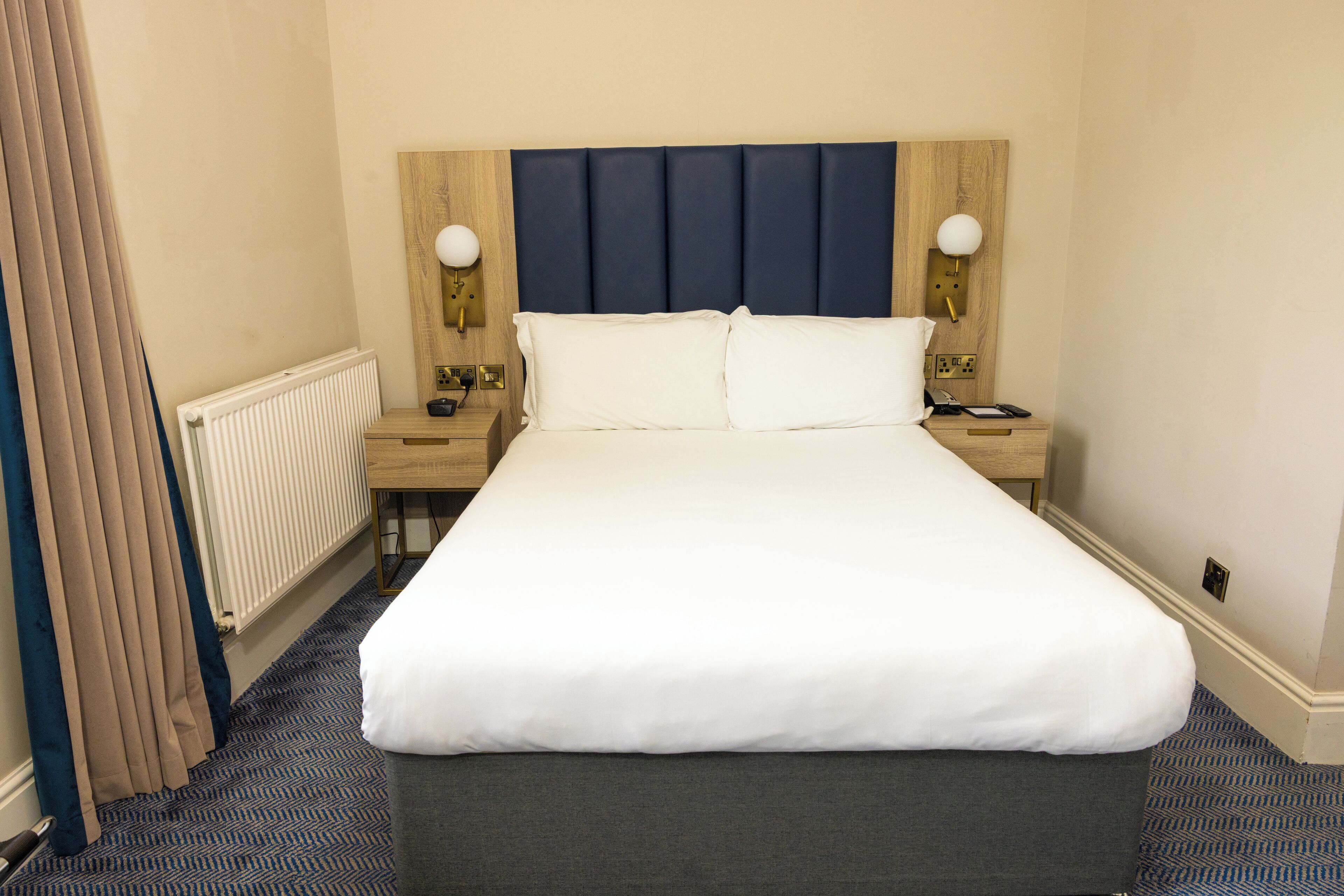 A hotel room with a large bed, a wooden headboard, two bedside tables with lamps, a radiator, and a window with curtains.