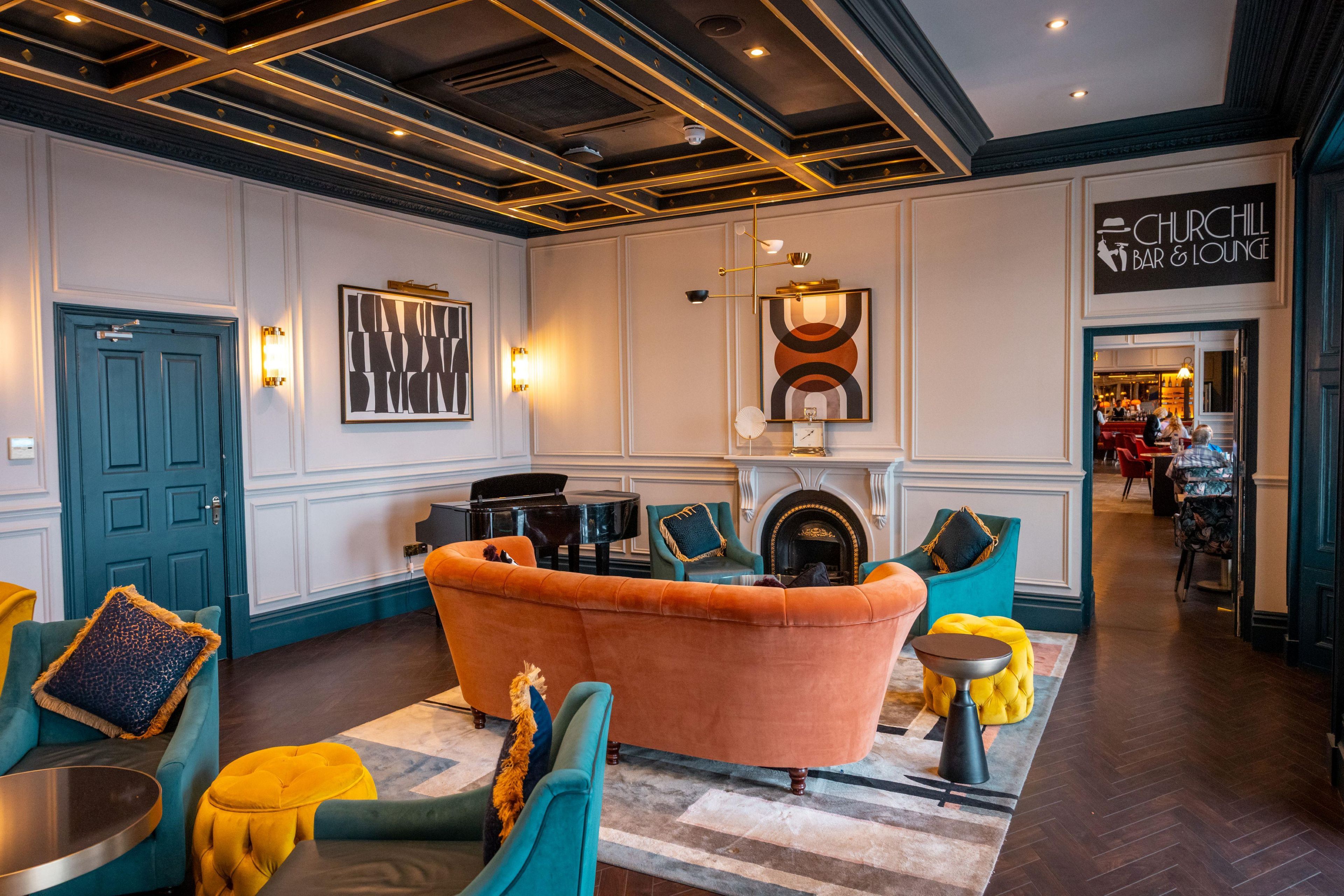 Eine Lounge mit einem Flügel, komfortabler Sitzgelegenheiten, einem Kamin und einem Schild für die Churchill Bar & Lounge.