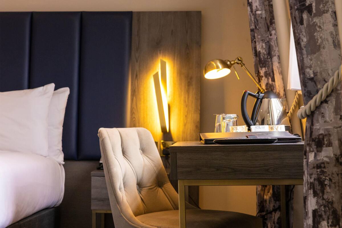 A hotel room with a bed, a chair, a desk with a lamp, and a window with curtains.