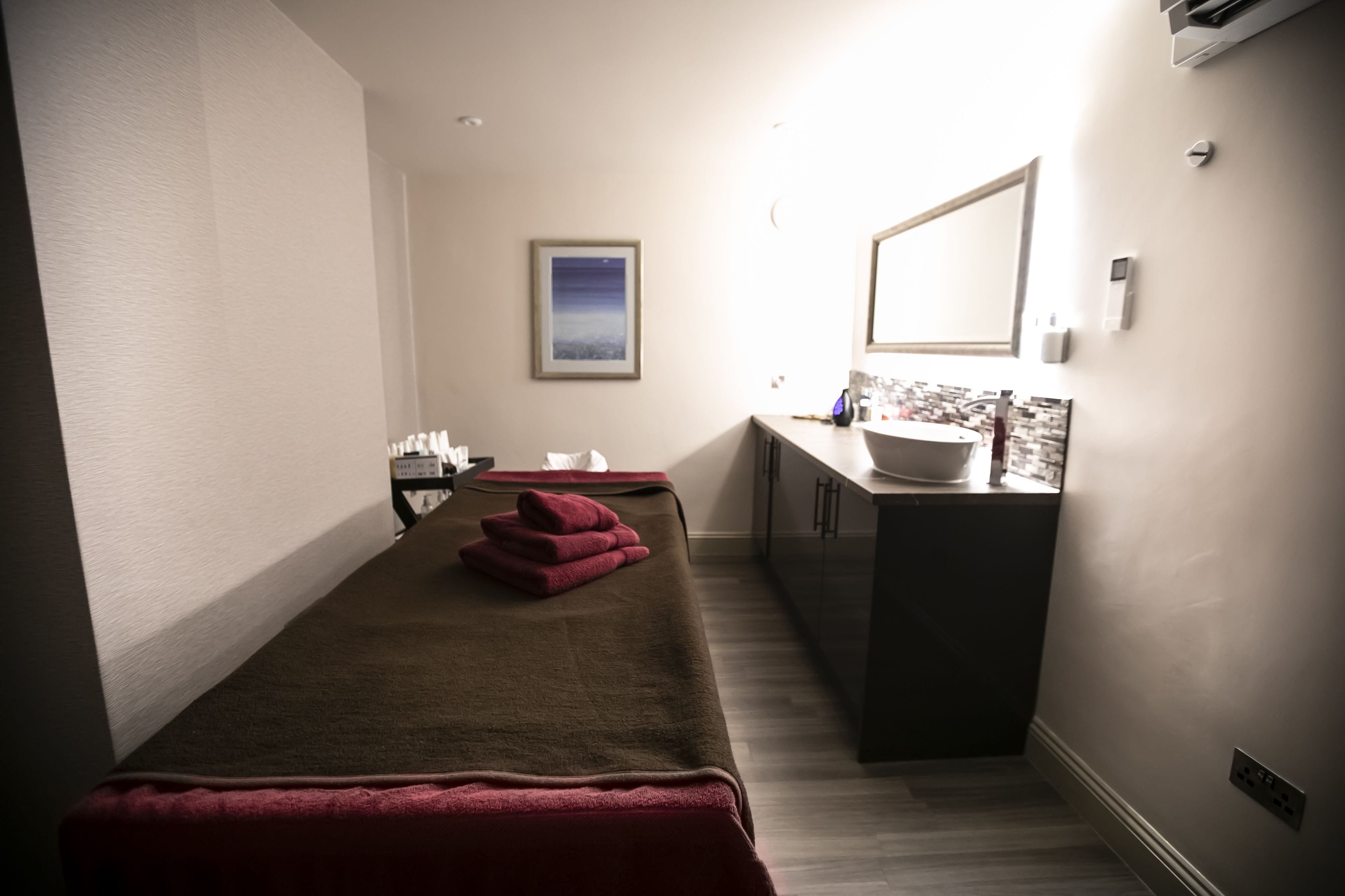 A spa room with a massage table, towels, a sink, and a framed picture on the wall.