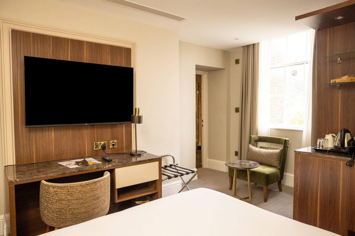 A hotel room with a flat-screen TV, a desk with a chair, a small table with a chair, and a cabinet.