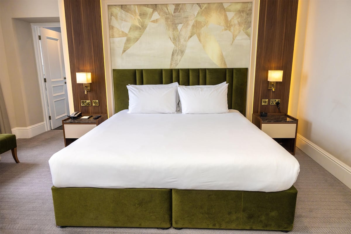 A hotel room with a large bed, two bedside tables, a green upholstered headboard, and a wall-mounted artwork.