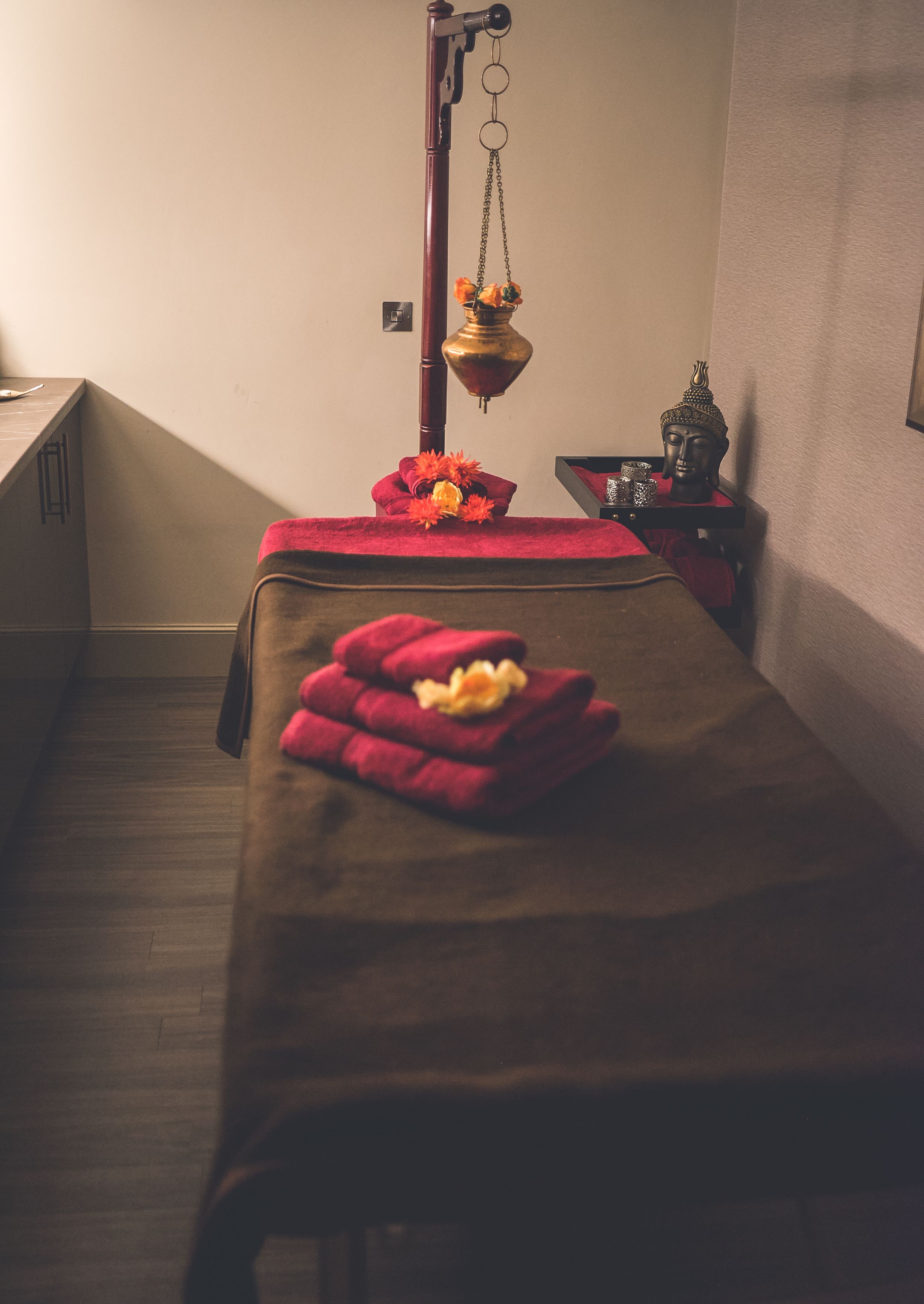 A massage table with red towels and flowers, a small Buddha statue, and a hanging brass lamp in a room with wooden flooring and a sloped ceiling.