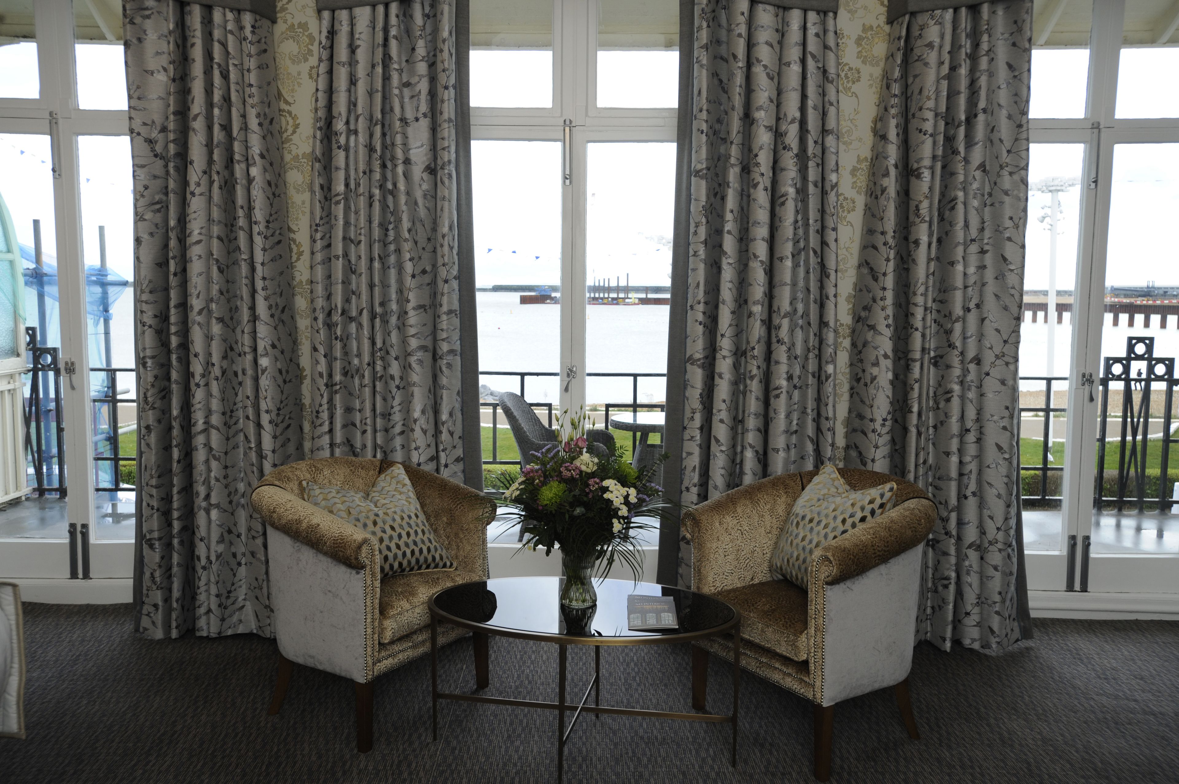 A room with two armchairs, a coffee table, and a vase with flowers. The room has large windows with curtains, a view of a body of water and a bridge.