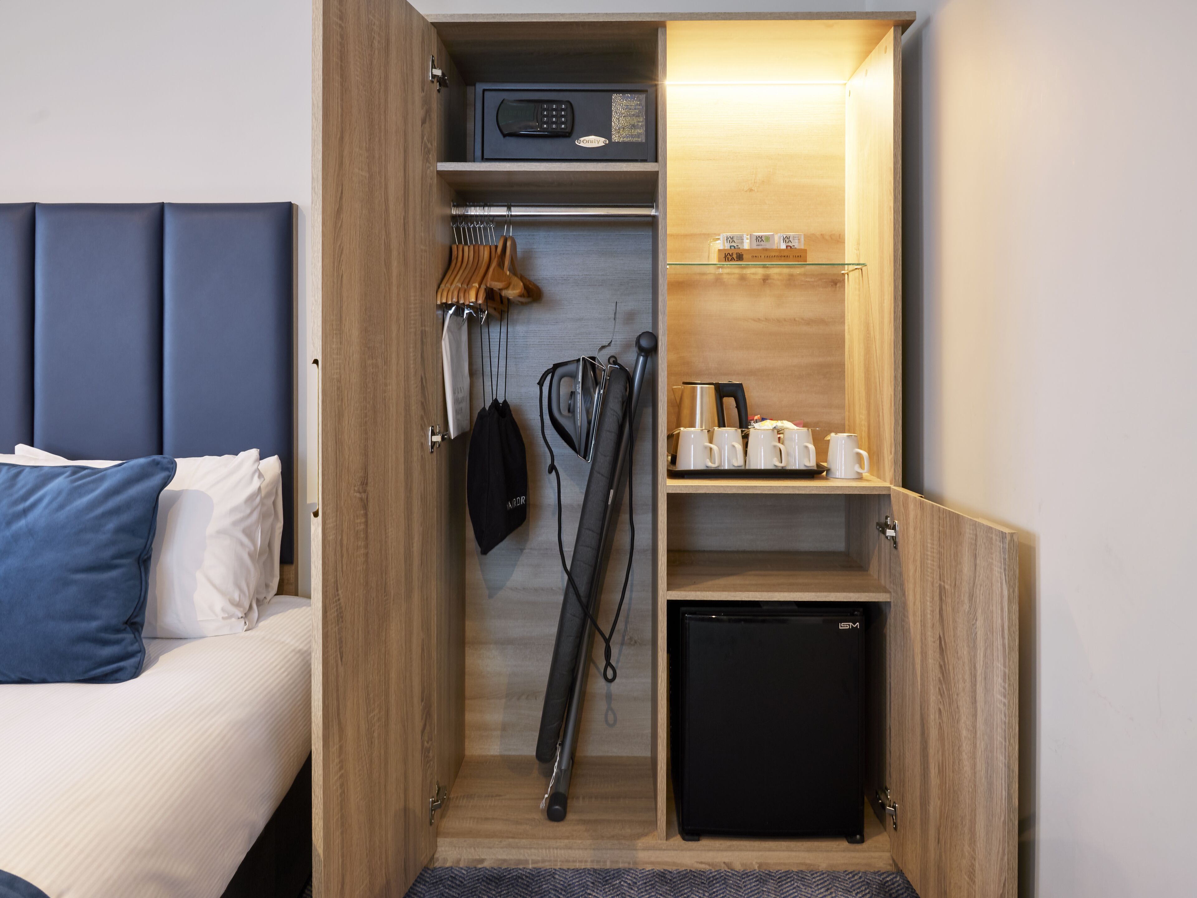 A compact hotel room with a bed, a small black refrigerator, a microwave, a kettle, and a set of mugs on a shelf.
