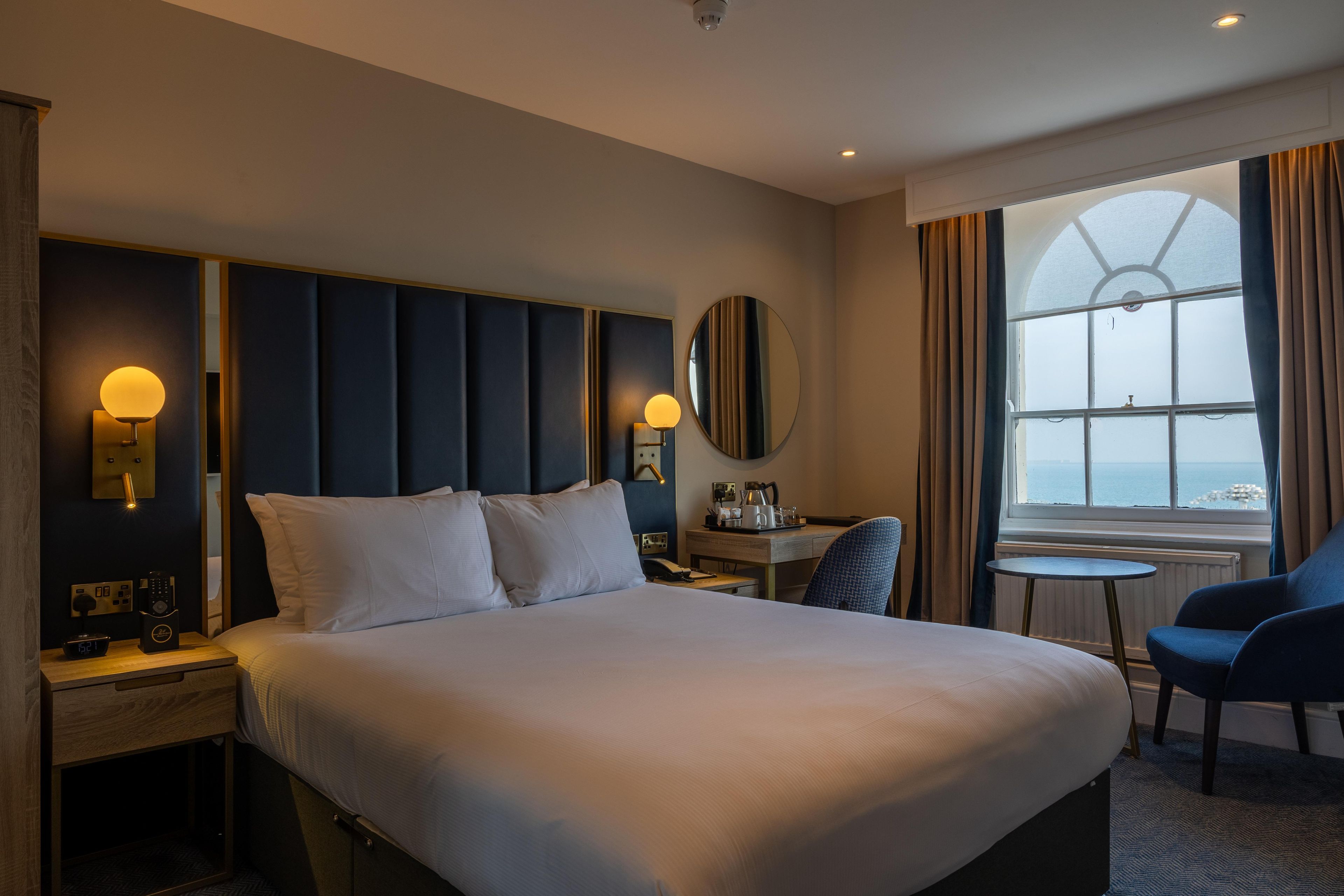 Habitación de hotel con una cama grande, lámparas de noche, un escritorio con silla y vista al mar por la ventana.