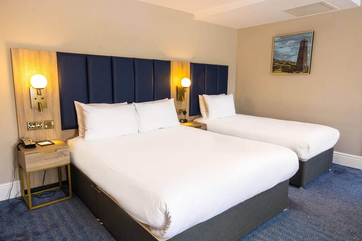 A hotel room with two beds, a wooden headboard, wall-mounted lamps, a painting, and a small bedside table with a phone.