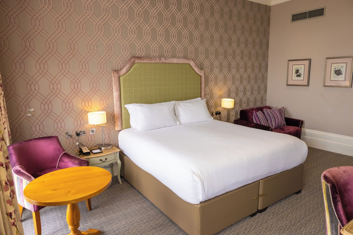 Una habitación de hotel con una cama grande, una mesita pequeña, una silla, una mesita de noche y una lámpara.