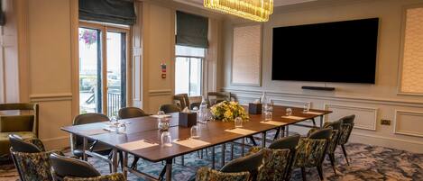 Une salle de conférence avec une longue table, des chaises, un lustre, un téléviseur à écran plat et une grande fenêtre.