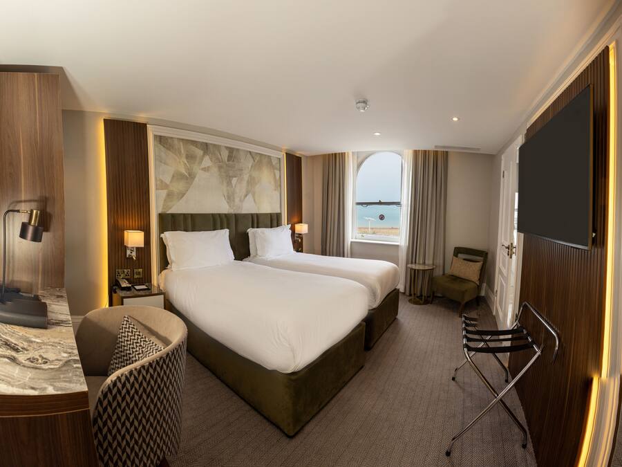 A hotel room with two beds, a desk, a chair, a TV, and a view of the beach.