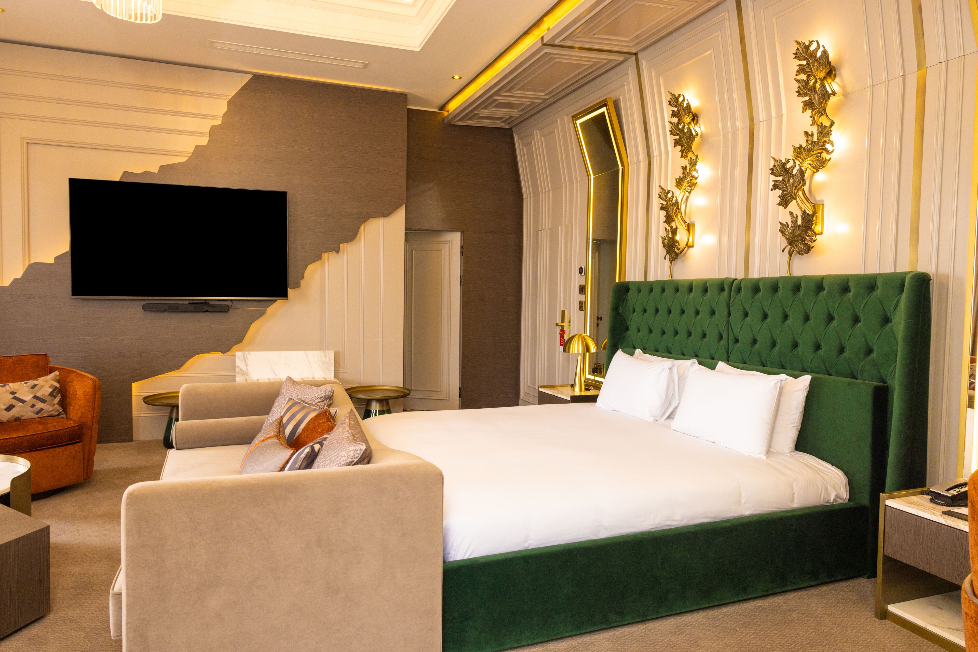 Una camera d'hotel con un letto grande, una televisione, una testiera imbottita verde e una zona salotto con un divano e una poltrona.