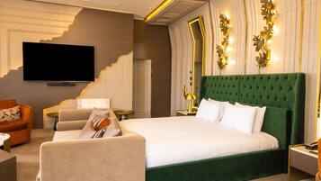 Una camera d'hotel con un letto grande, una televisione, una testiera imbottita verde e una zona salotto con un divano e una poltrona.