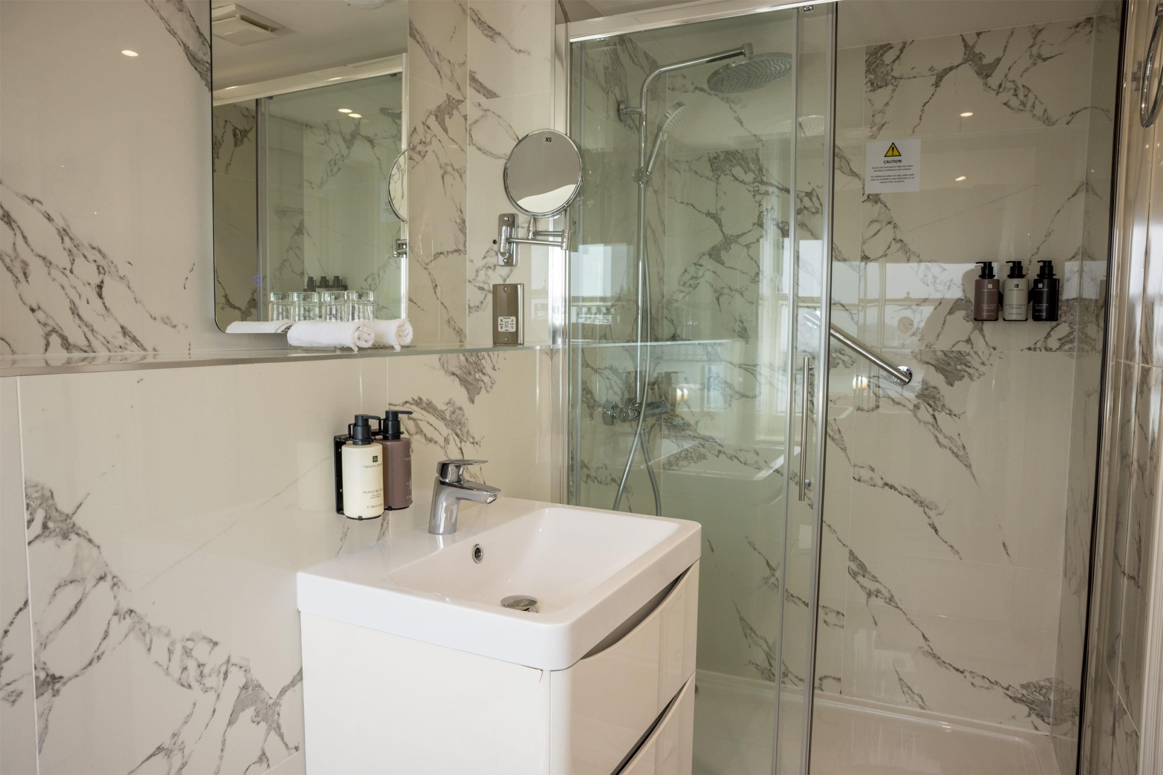 A bathroom with a marble wall, a glass shower enclosure, a white sink, and toiletries on the countertop.