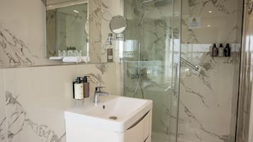 Un baño con pared de mármol, cabina de ducha de vidrio, lavamanos blanco y artículos de aseo sobre la encimera.
