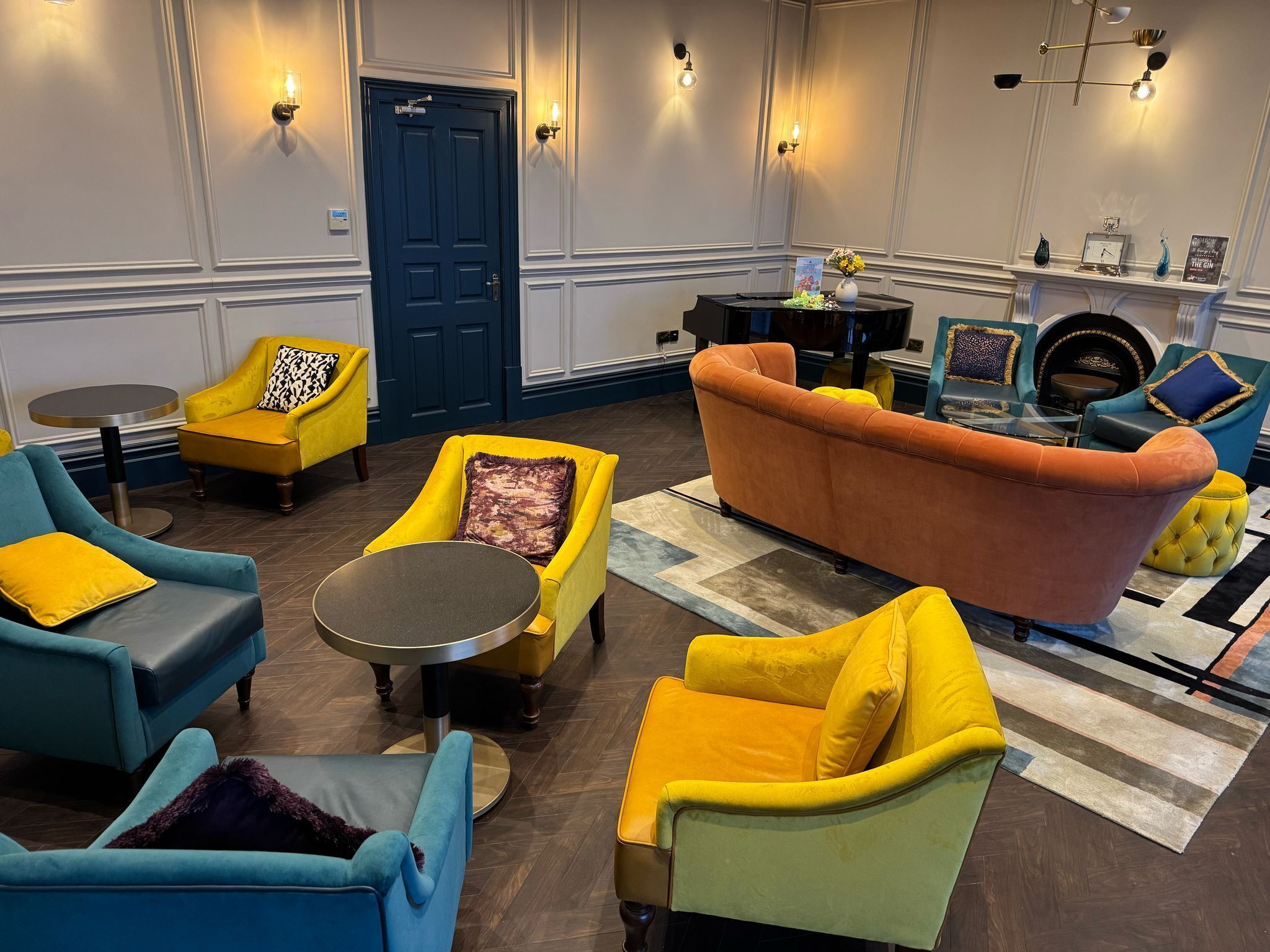 Un área de descanso moderna con una puerta azul, sillones azul y amarillo, una mesa redonda para café y una chimenea decorada.