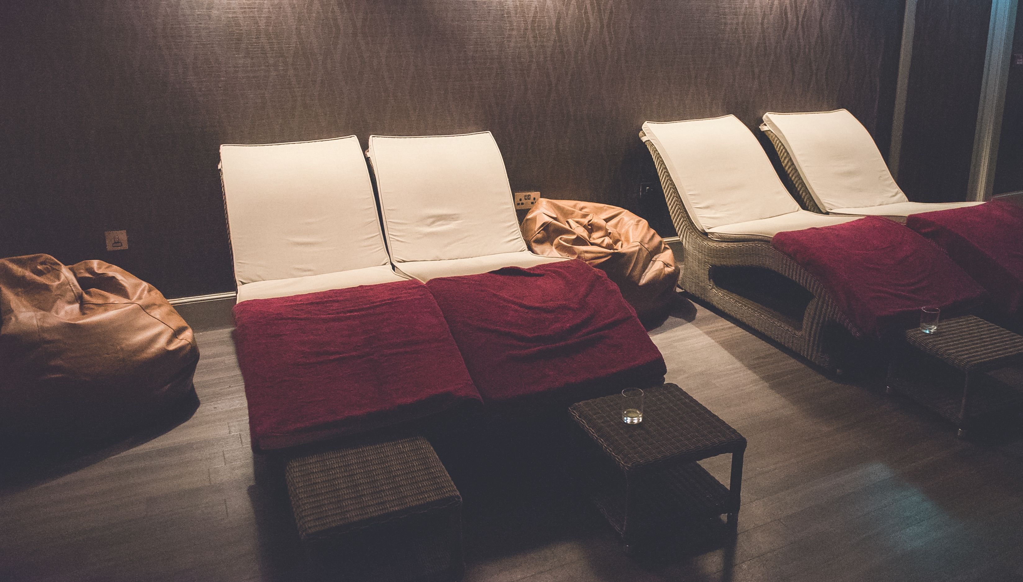 Una habitación con dos sillones reclinables, cada uno con una almohada blanca y una manta roja. Hay una silla de mimbre con un cojín rojo y una mesita con un vaso y un cuenco. Una bolsa marrón está en el suelo.