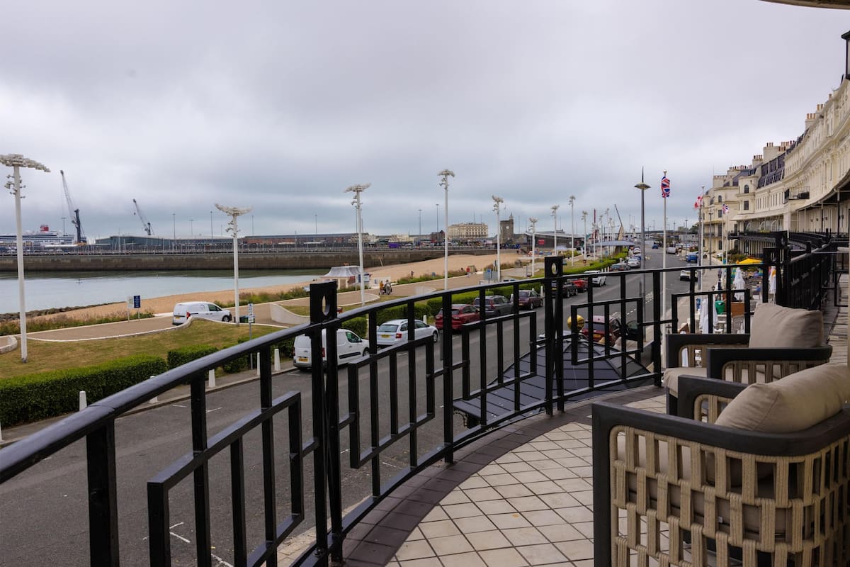 A balcony with a view of a waterfront promenade, a promenade with street lamps, and a building with a clock tower in the background.