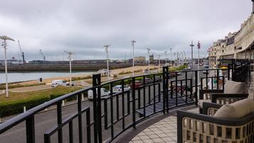 A balcony with a view of a waterfront promenade, a promenade with street lamps, and a building with a clock tower in the background.