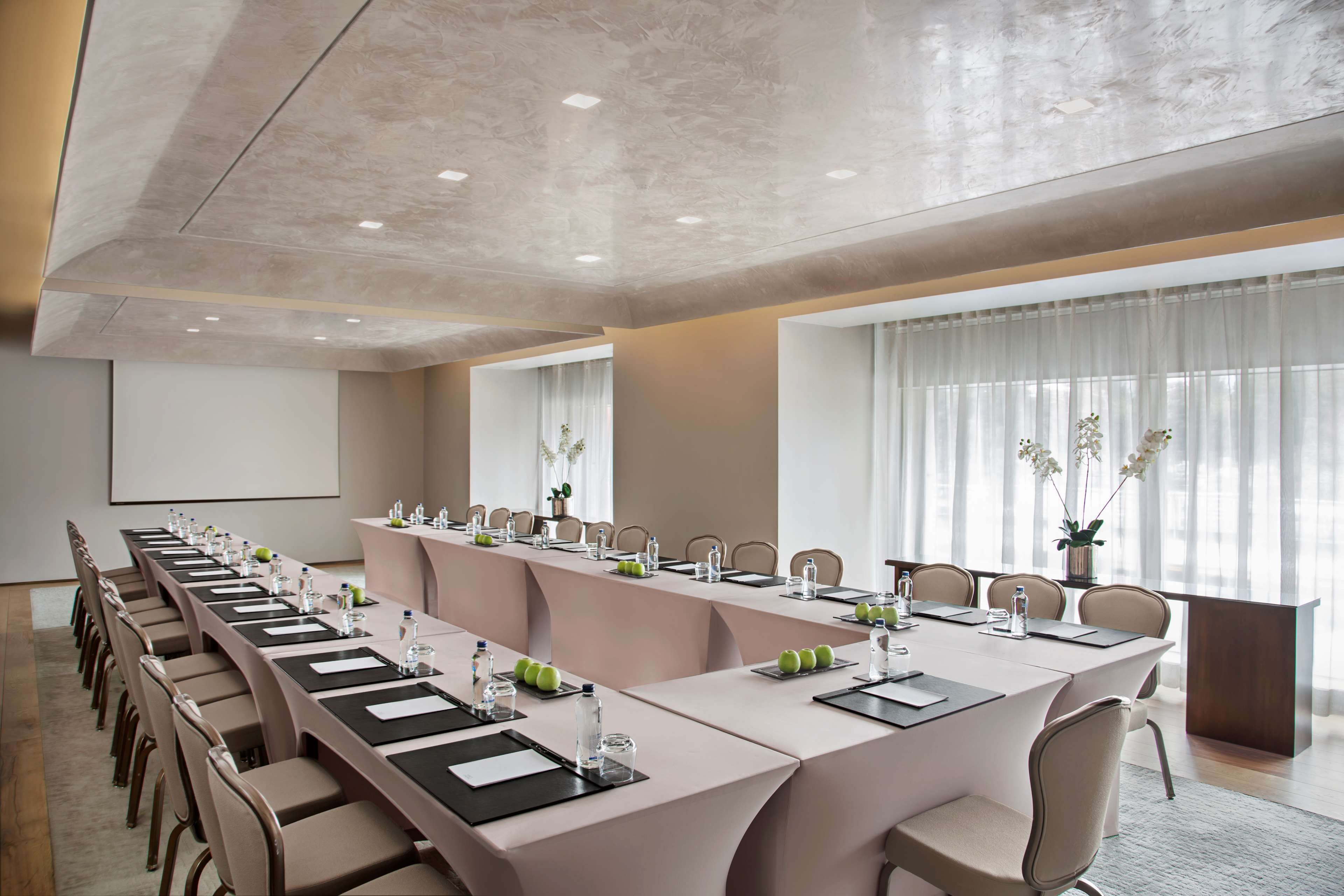 A conference room with a long table, chairs, a projector screen, and a large window with curtains.