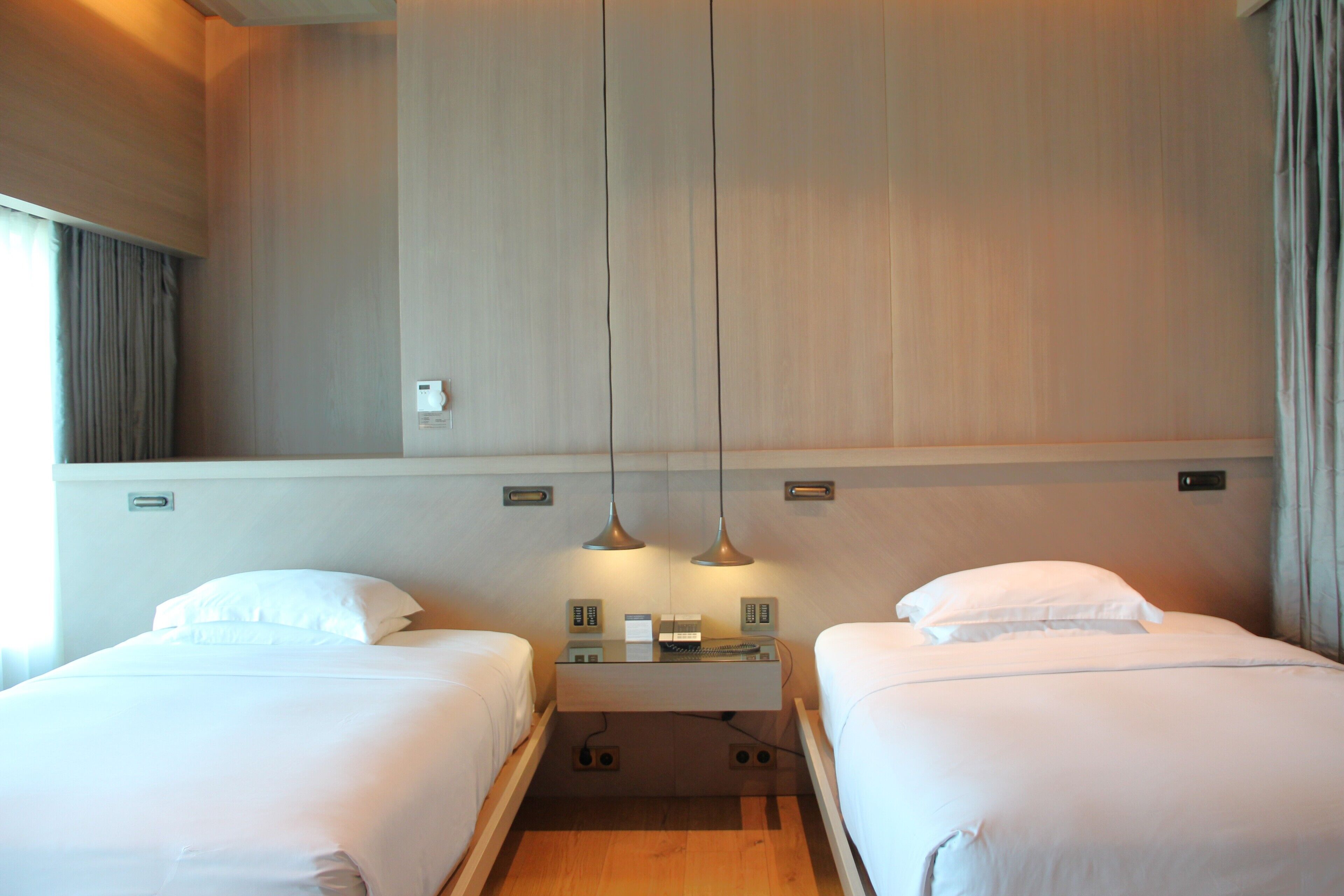A hotel room with two beds, a bedside table, a lamp, and a wall-mounted control panel.