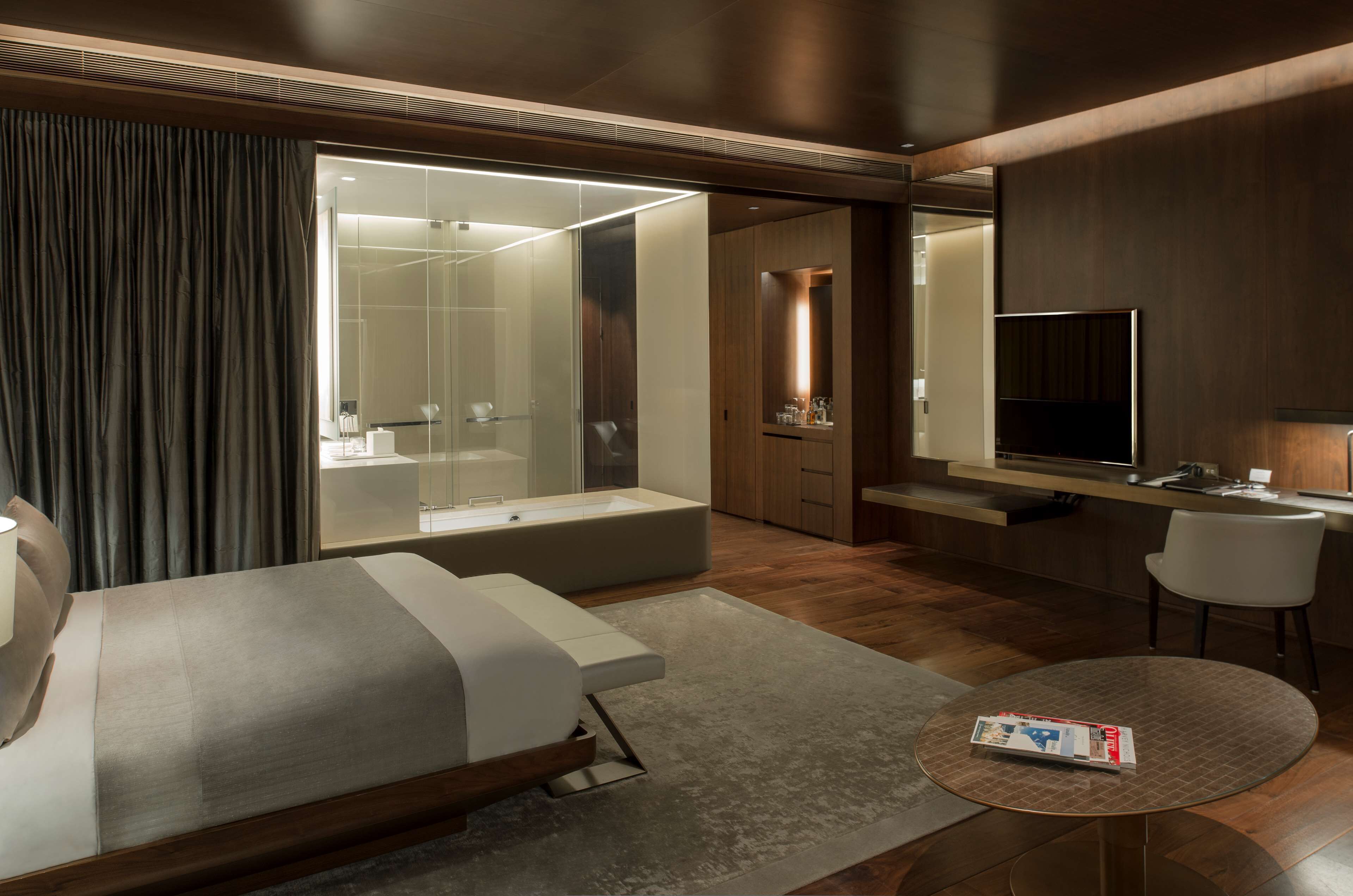 A modern hotel room with a bed, a round table, a TV, a bathroom with a glass shower, and wooden paneling.