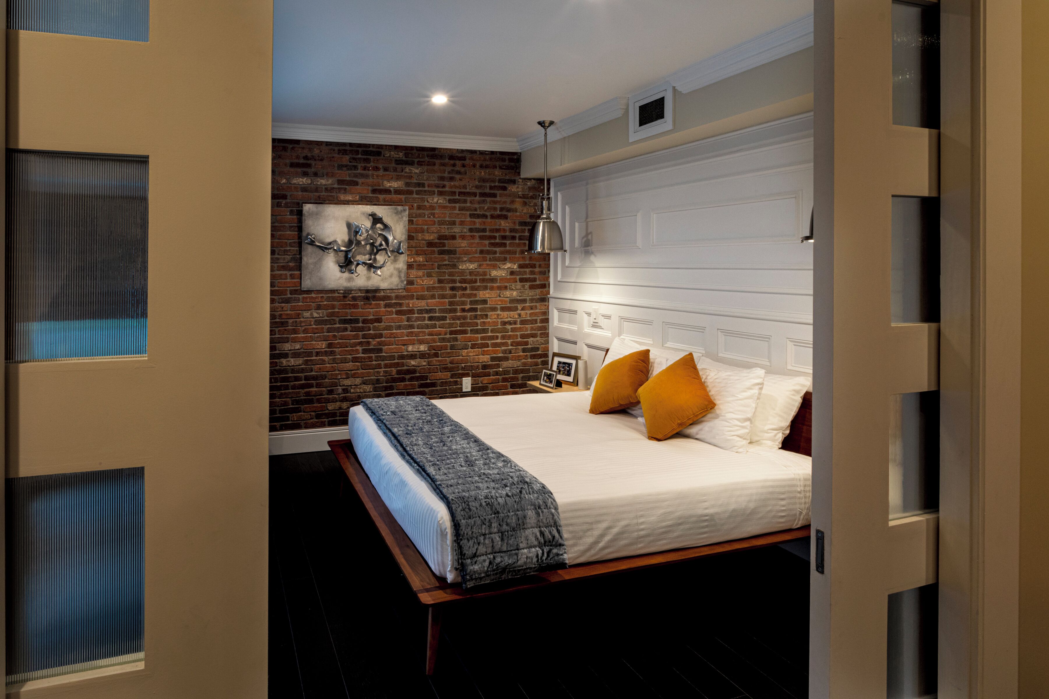 A bedroom with a brick wall, a bed with white bedding and orange pillows, a gray throw blanket, and a hanging lamp.